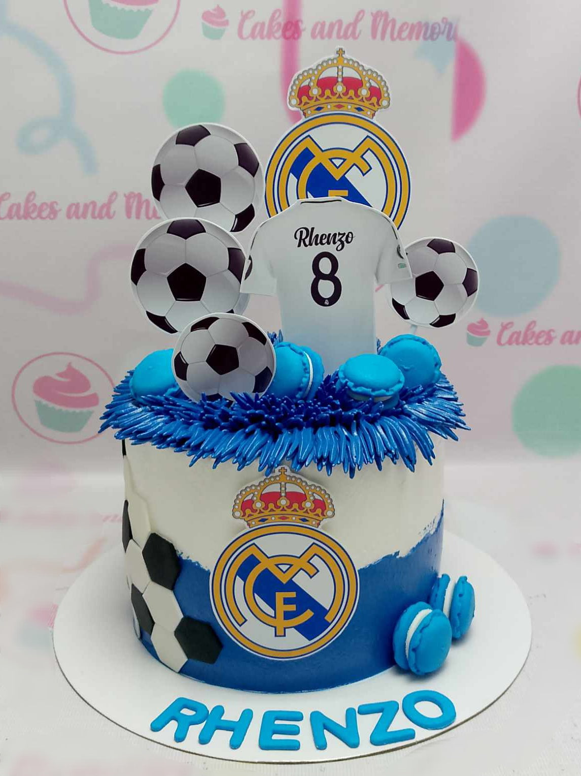 This custom soccer cake features a sleek white and blue design, adorned with a soccer ball and a Real Madrid football jersey for Rhenzo's 8th birthday. Completed with macarons and printout toppers, it captures the essence of football sports and Cristiano Ronaldo.