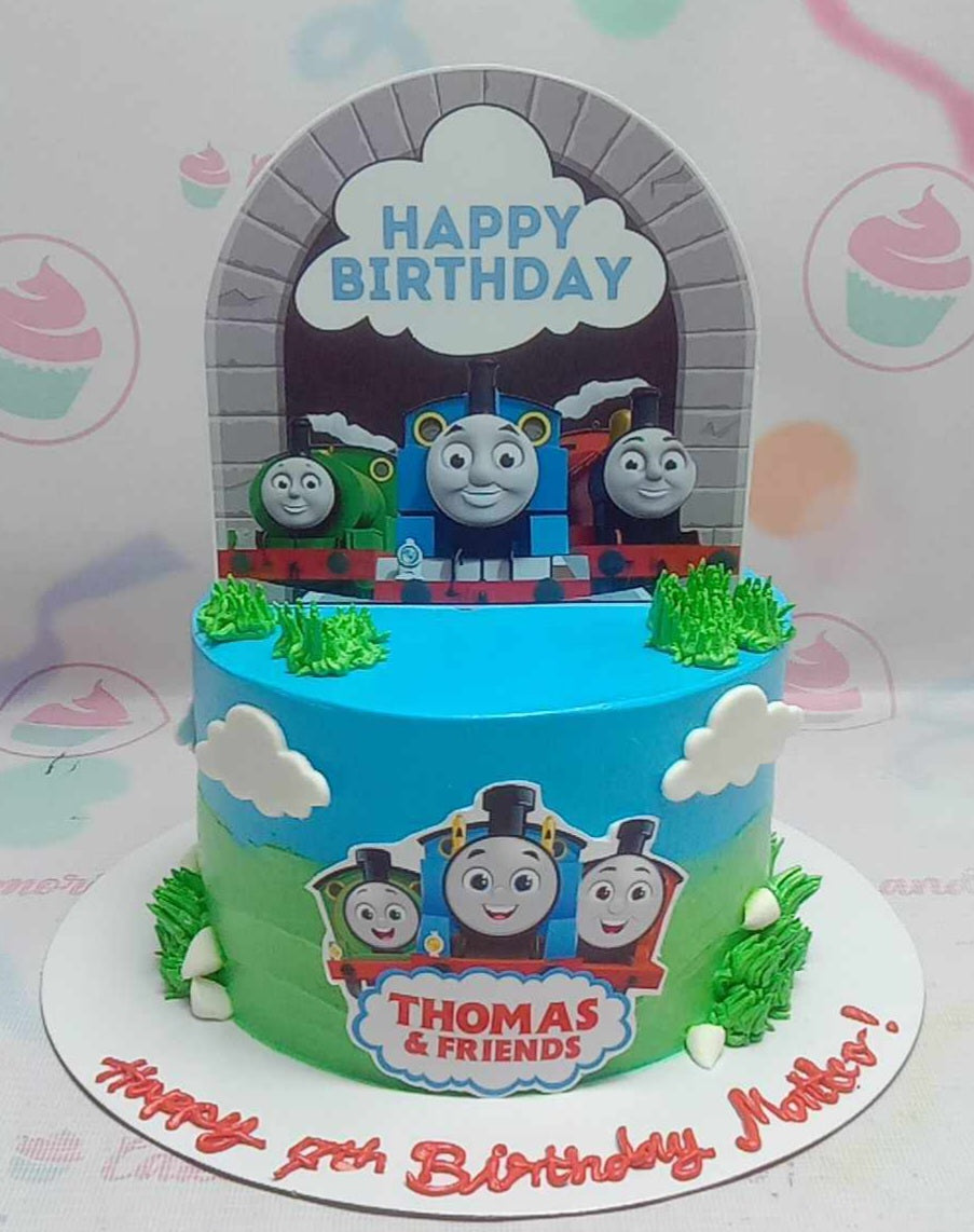 A cheerful blue and green Bus Train Trucks Cake featuring Thomas the Train, Percy, and James the Red Engine, perfect for a 4th birthday celebration. Crafted as a single tier with delightful printout toppers, this customized cake is ideal for young train enthusiasts.