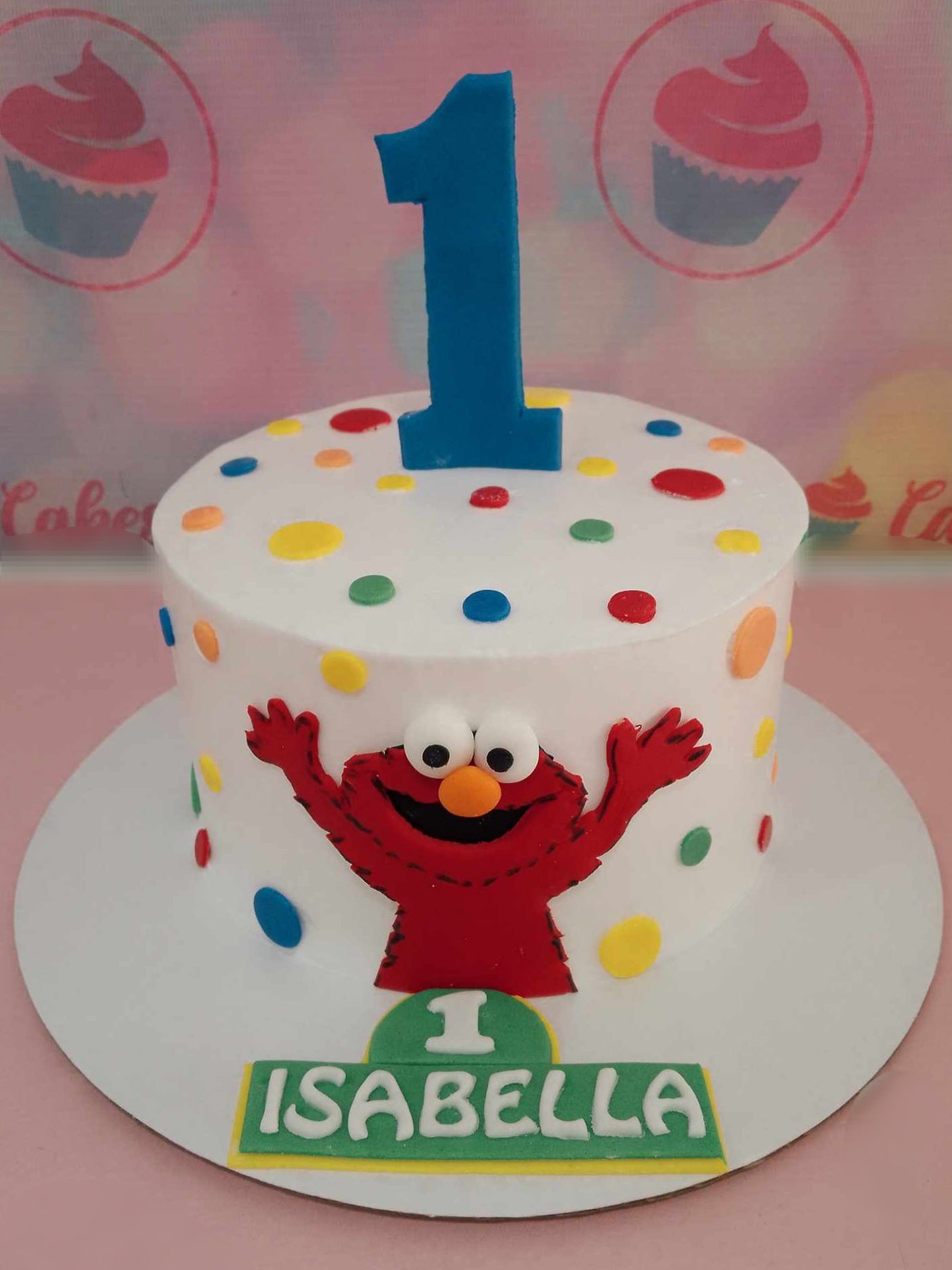 Celebrate Isabella's 1st birthday with a vibrant Sesame St Cake, featuring colorful dots and delightful printout toppers of Elmo, Big Bird, and Cookie Monster. This whimsical one-tier design, adorned with white icing, is the perfect centerpiece for a festive party!