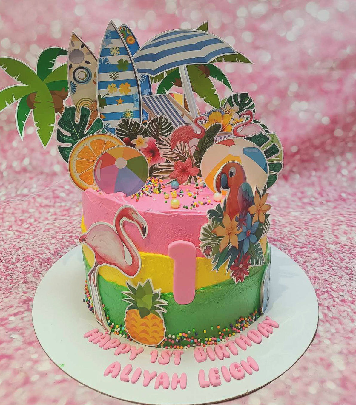 A stunning tropical cake, adorned with pink icing and hints of green and yellow, features adorable flamingos, a parrot, and cheerful beach-themed elements like surfboards and a beach ball. Celebrating Aliyah Leigh's 1st birthday, this Hawaiian-inspired creation is perfect for summer festivities, complete with delightful printout toppers and a pineapple garnish.