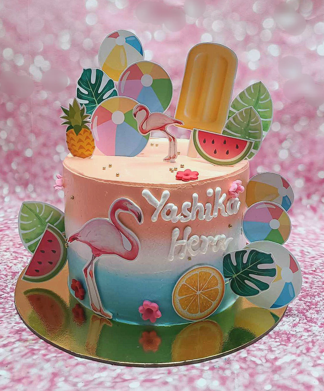 This tropical cake showcases a stunning pink and blue ombre design, adorned with playful beach balls, a flamingo, and fresh fruit accents including pineapple, watermelon, and lemon slices. Perfect for a summer beach outing, it features fun printout toppers and a touch of gold with popsicle ice cream decor.