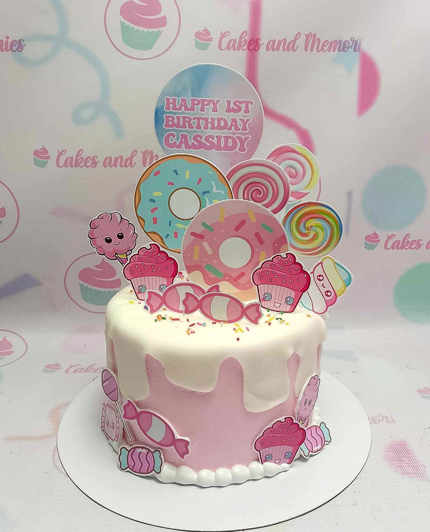 This enchanting Candyland Cake showcases a delightful pink design, perfect for Cassidy's 1st birthday celebration. Adorned with colorful donuts, lollipops, and candies, this one-tier cake features charming printout toppers for a festive touch.