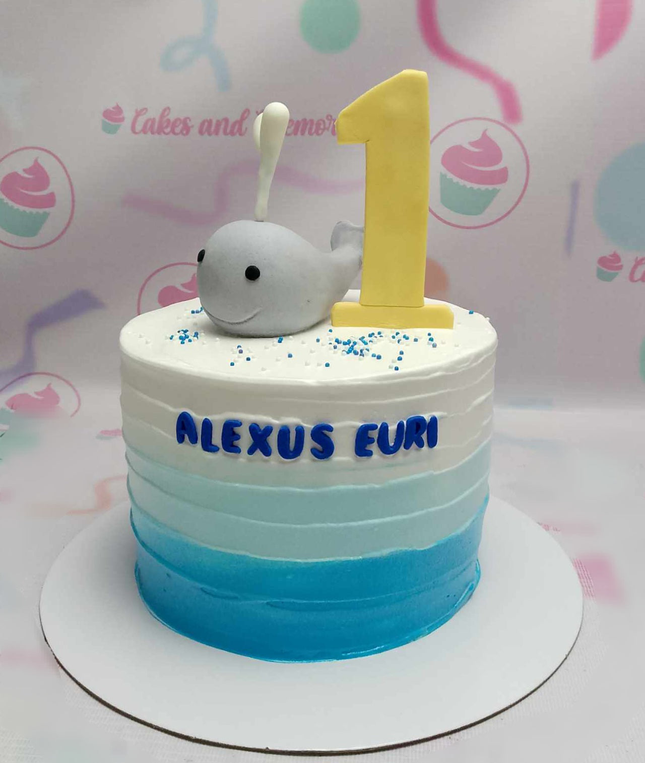 This Under the Sea Cake features a stunning blue ombre design with a cute whale and "1" on top, adorned with colorful sprinkles. Perfect for celebrating Alexus Euri's first birthday, this one-tiered cake is a delightful centerpiece for a baby boy's special day.