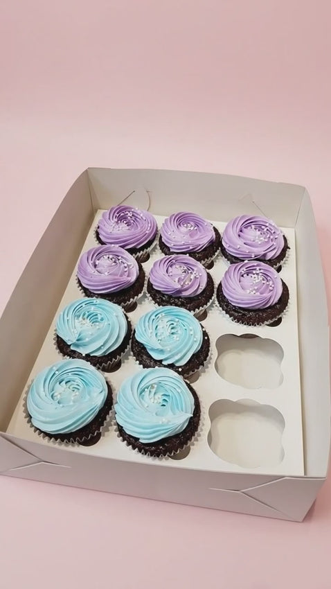 A dozen cupcakes with six pastel blue and six lavender swirled icing tops decorated with white sprinkles in a white box.