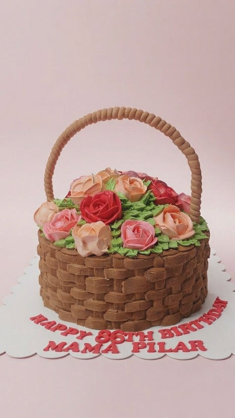 A round 1-tier cake designed as a woven brown basket overflowing with pink and red buttercream roses, with a piped basket handle and the message Happy 86th birthday Mama Pilar on the board.