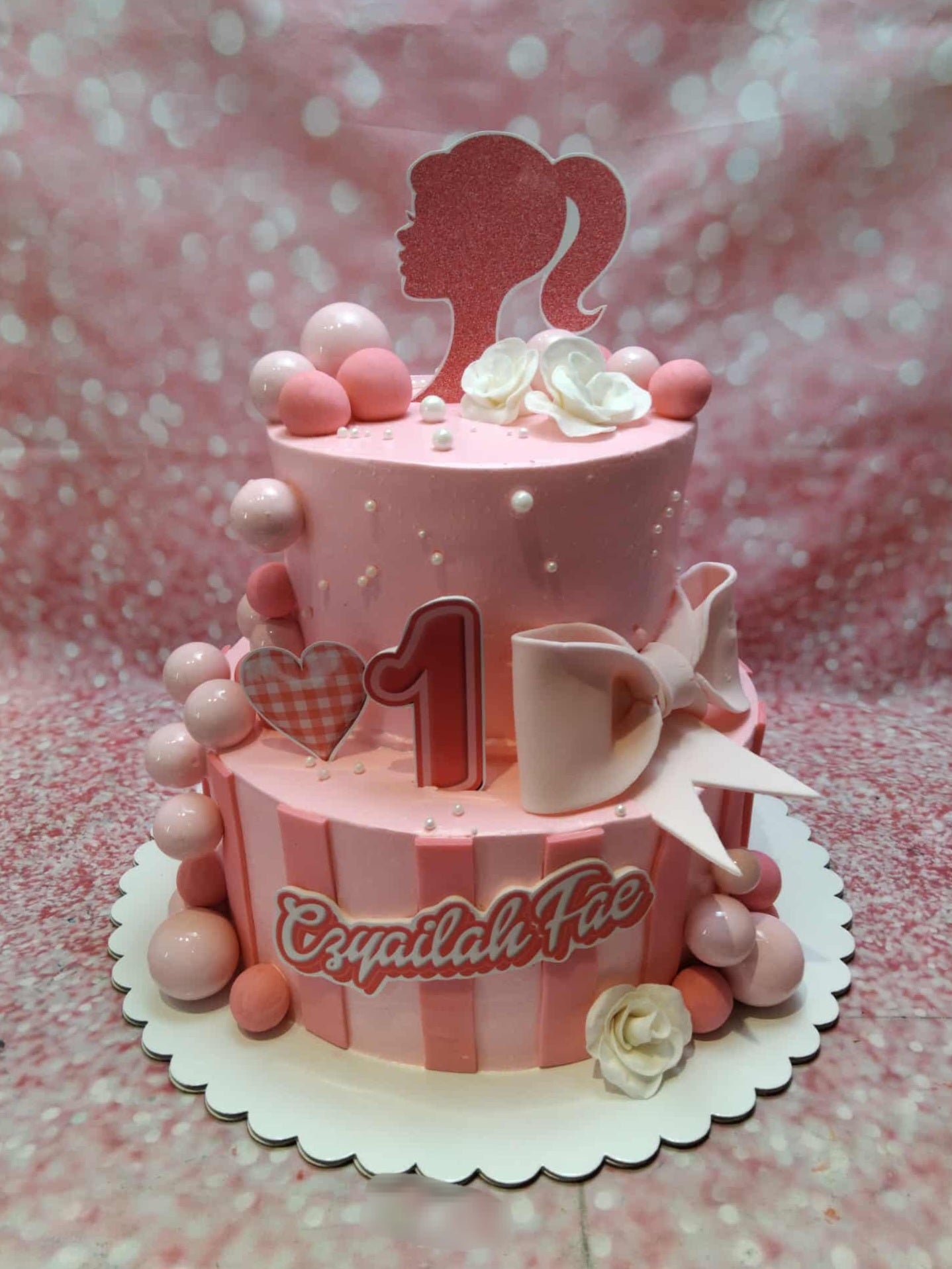Two-tier pink Barbie 1st birthday cake with a glittery silhouette, number one, name Czyailah Fae, and pink stripes.