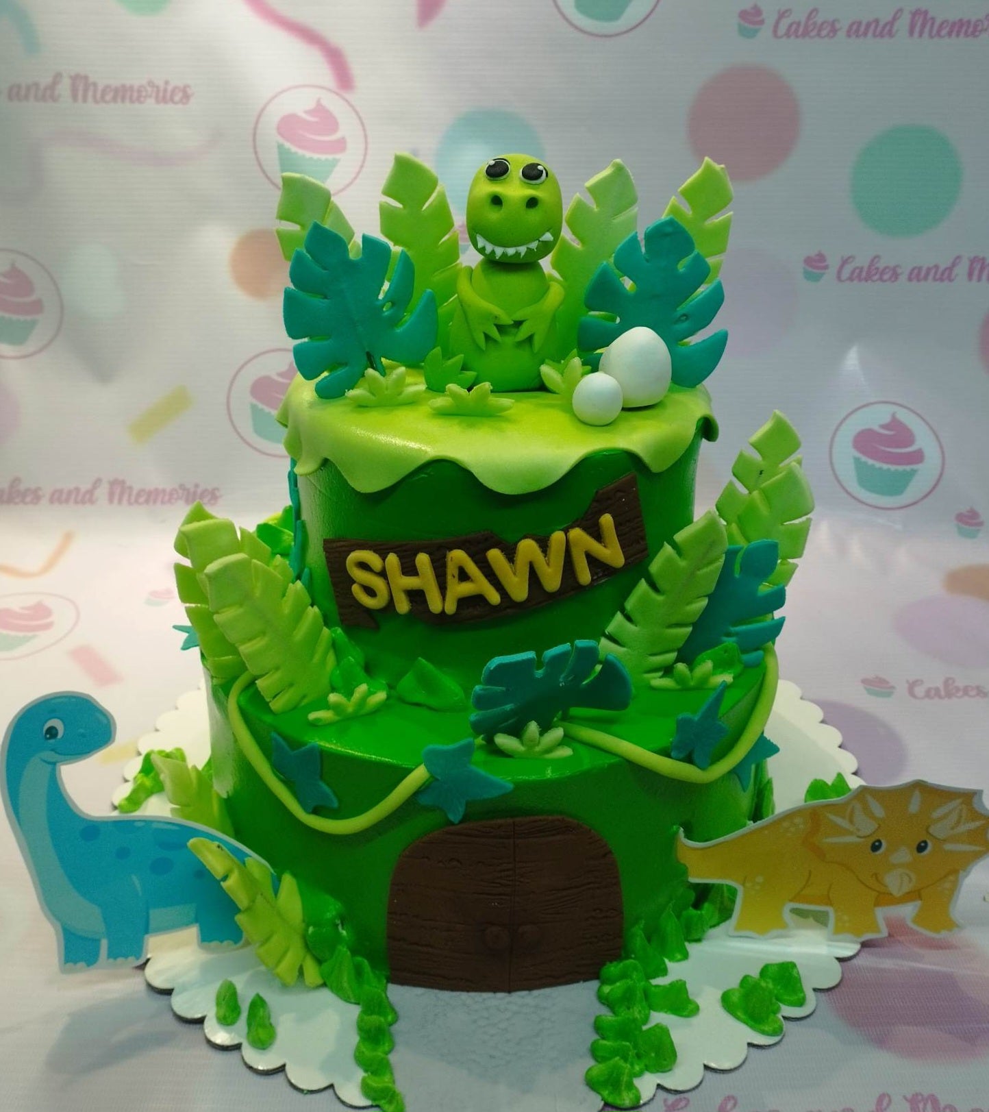 This custom decorated cake features dinosaurs on a blue background. The cake includes a brontosaurus, triceratops, and a raptor in green, perfect for a dinosaur-loving kid's birthday. Bring Jurassic Park and Jurassic World to life with this dinosaur-themed cake.
